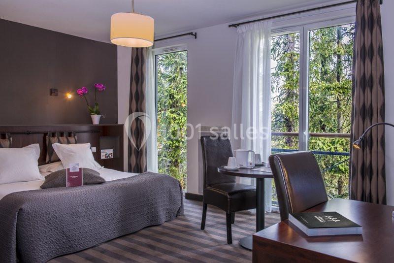 Chambre d'hôtel lumineuse avec lit double, table avec chaises, bureau, orchidée et grandes fenêtres donnant sur des arbres.