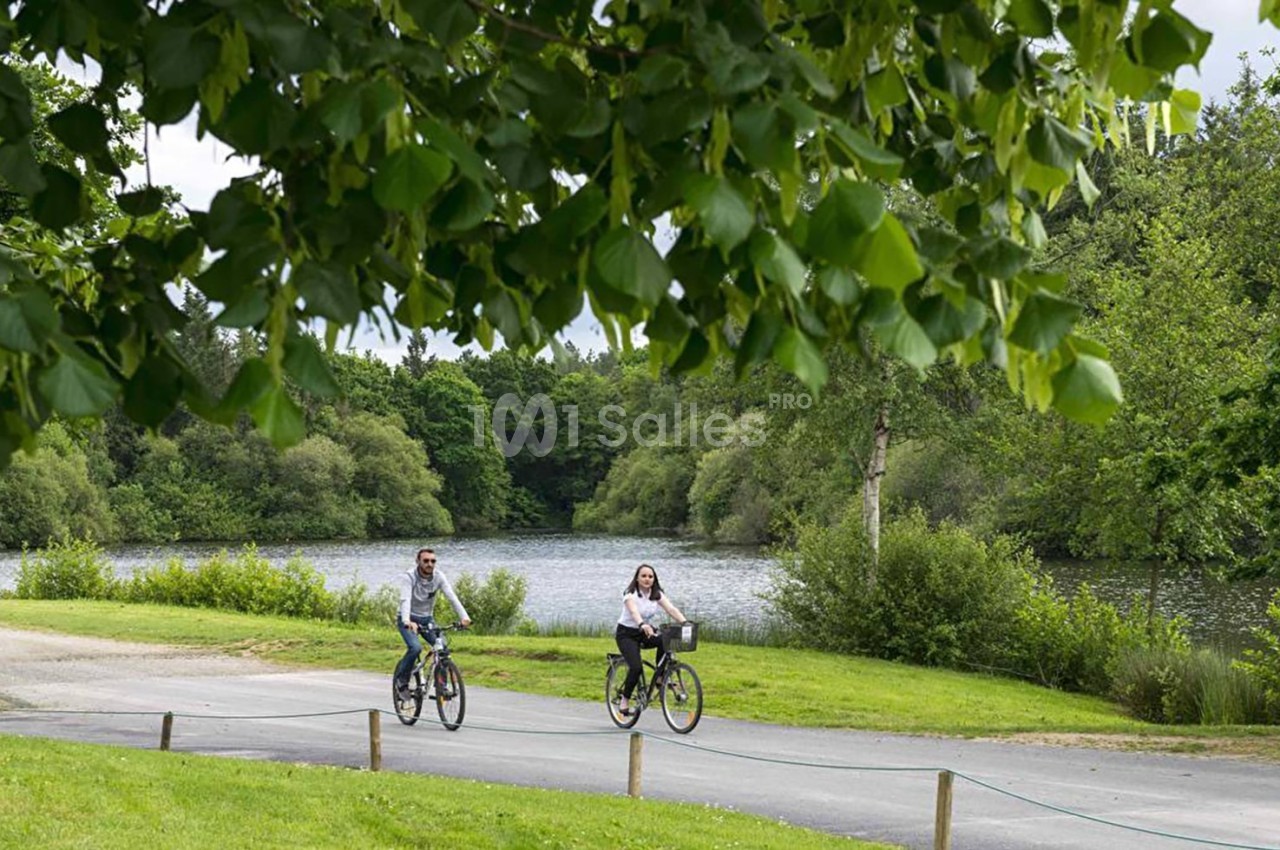 Deux personnes font du vélo sur un chemin bordé d'arbres, avec un lac et une végétation dense en arrière-plan.
