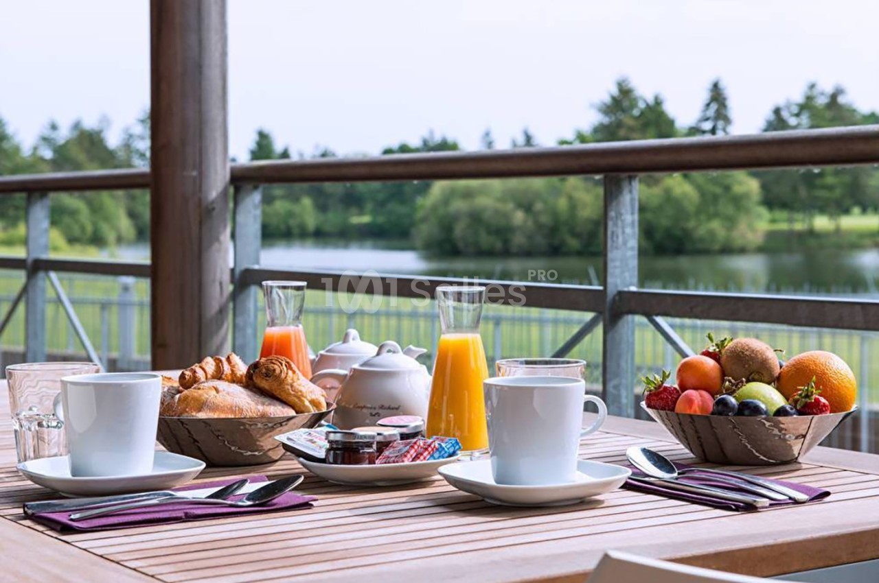 Petit-déjeuner servi en terrasse avec croissants, fruits, jus d'orange et vue sur un lac entouré de verdure.