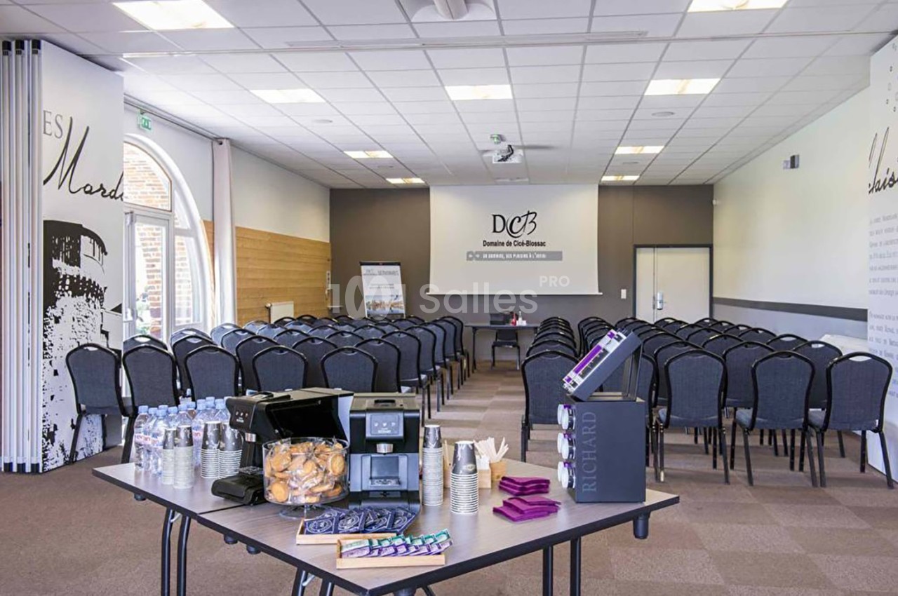 Salle de conférence aménagée avec des rangées de chaises, une table de collation au premier plan et un écran au fond.
