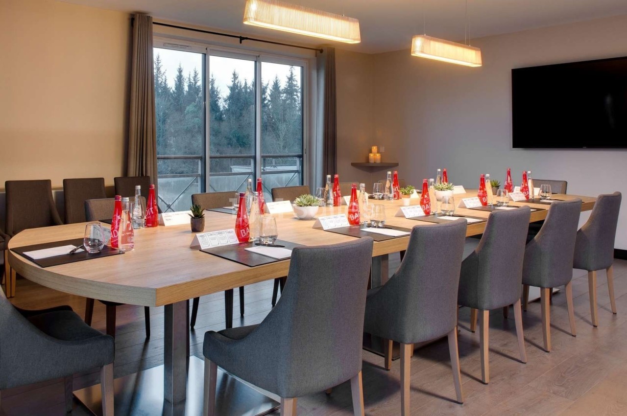Salle de réunion lumineuse avec une grande table ovale, chaises grises, écran mural et vue sur l'extérieur.