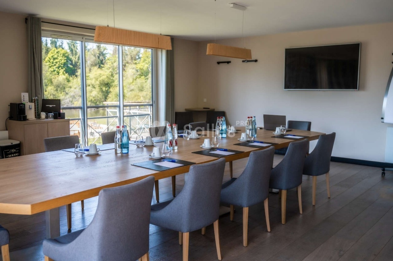 Salle de réunion lumineuse avec une grande table en bois, chaises grises, écran mural et vue sur l'extérieur verdoyant.