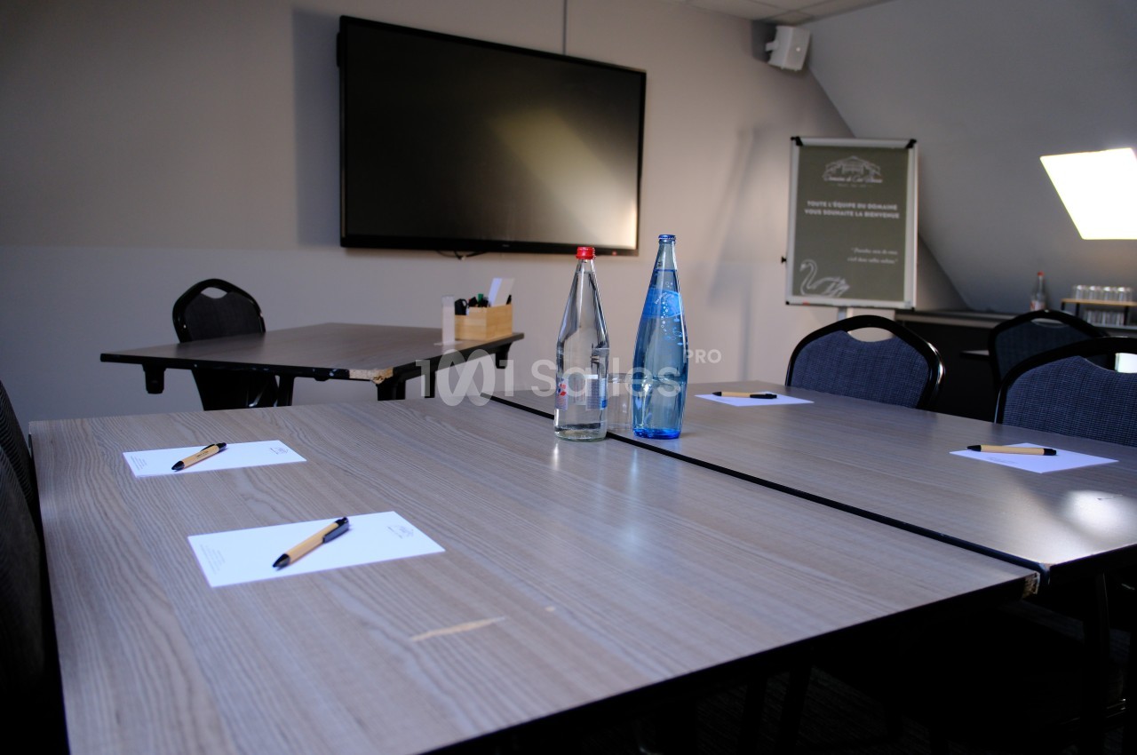 Salle de réunion avec tables en bois, bouteilles d'eau, stylos et blocs-notes, écran mural en arrière-plan.
