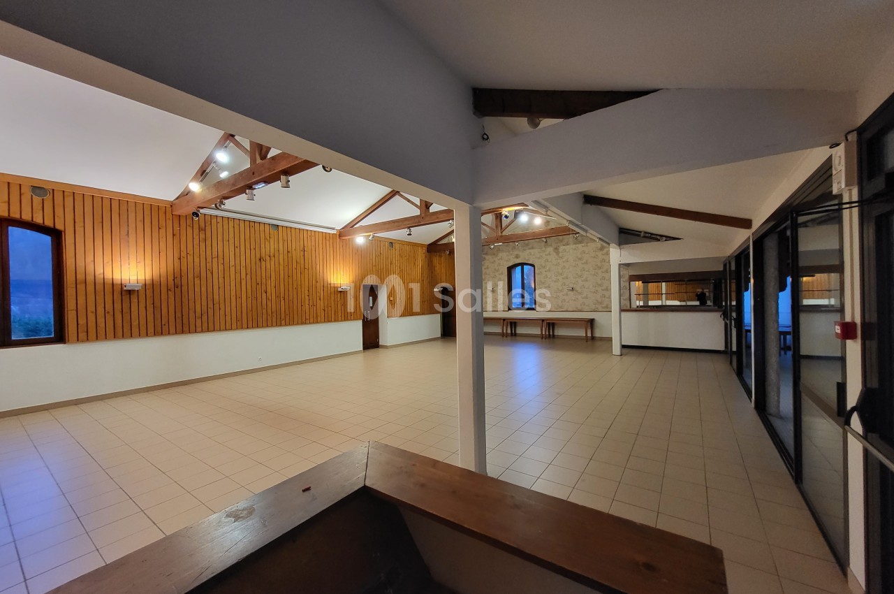 Salle spacieuse avec murs en bois et en pierre, carrelage clair, poutres apparentes et grandes fenêtres.