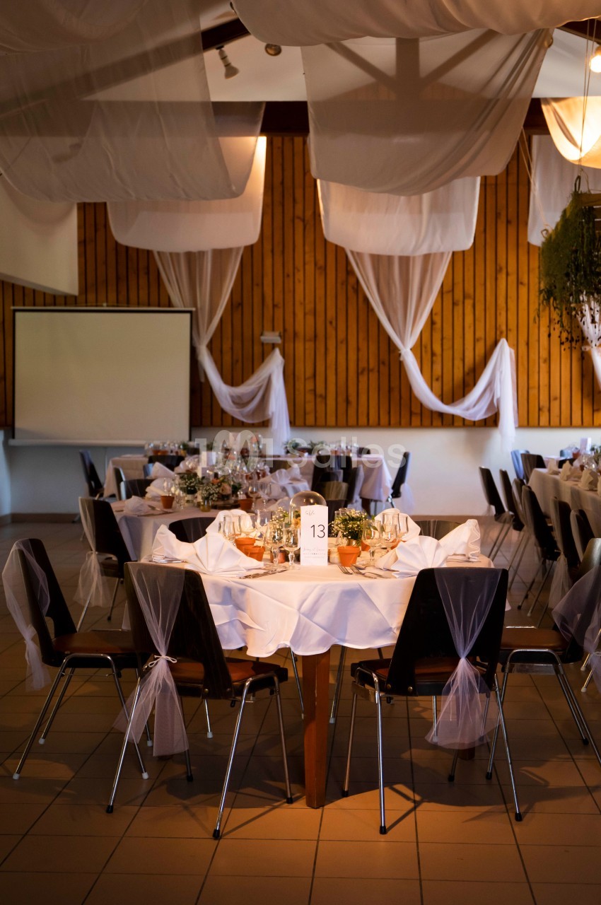 Salle de réception décorée avec des tables rondes, nappes blanches, chaises ornées de voiles et éclairage tamisé.