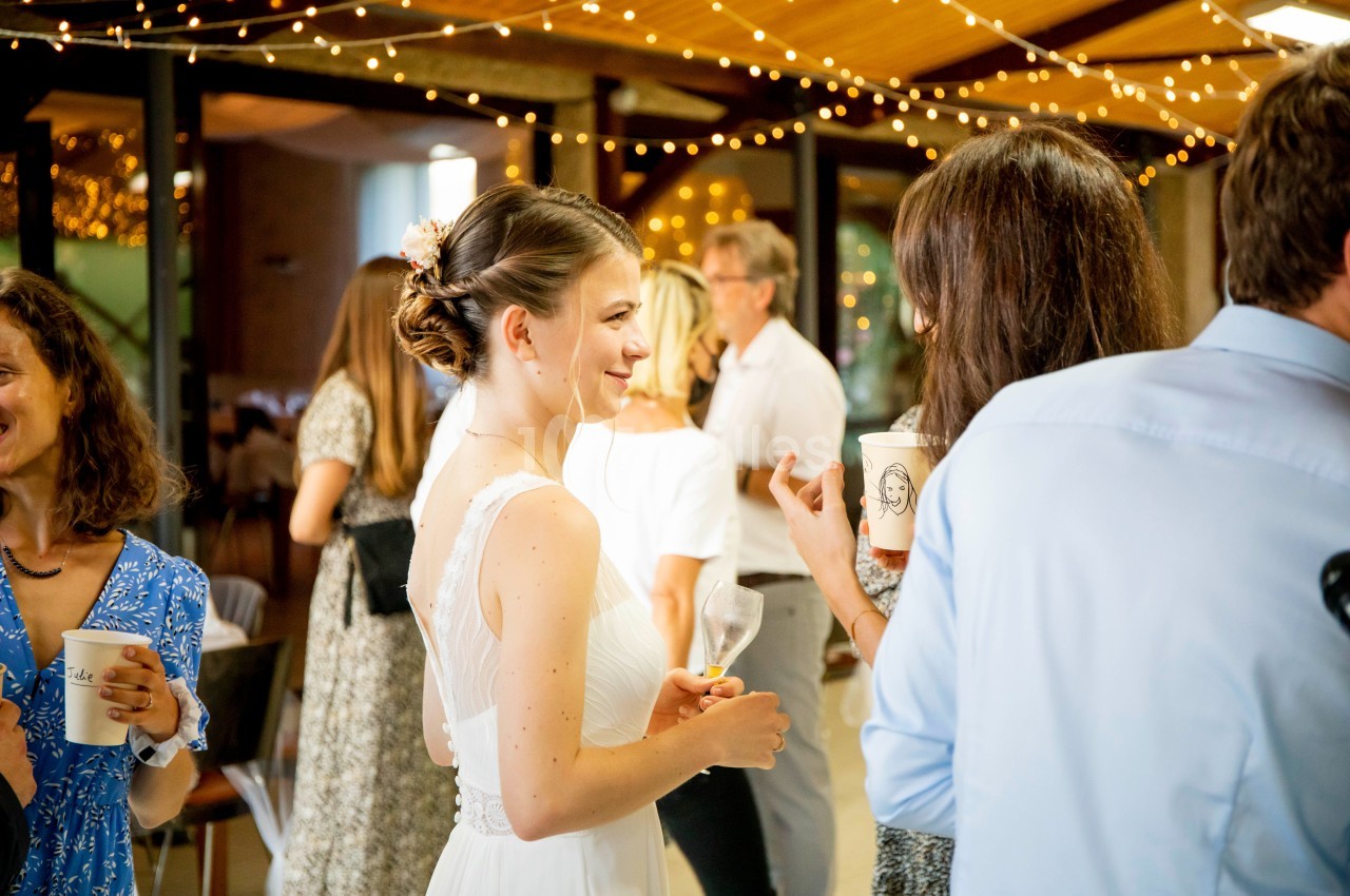 Une femme en robe blanche discute avec des invités dans une salle décorée de guirlandes lumineuses.