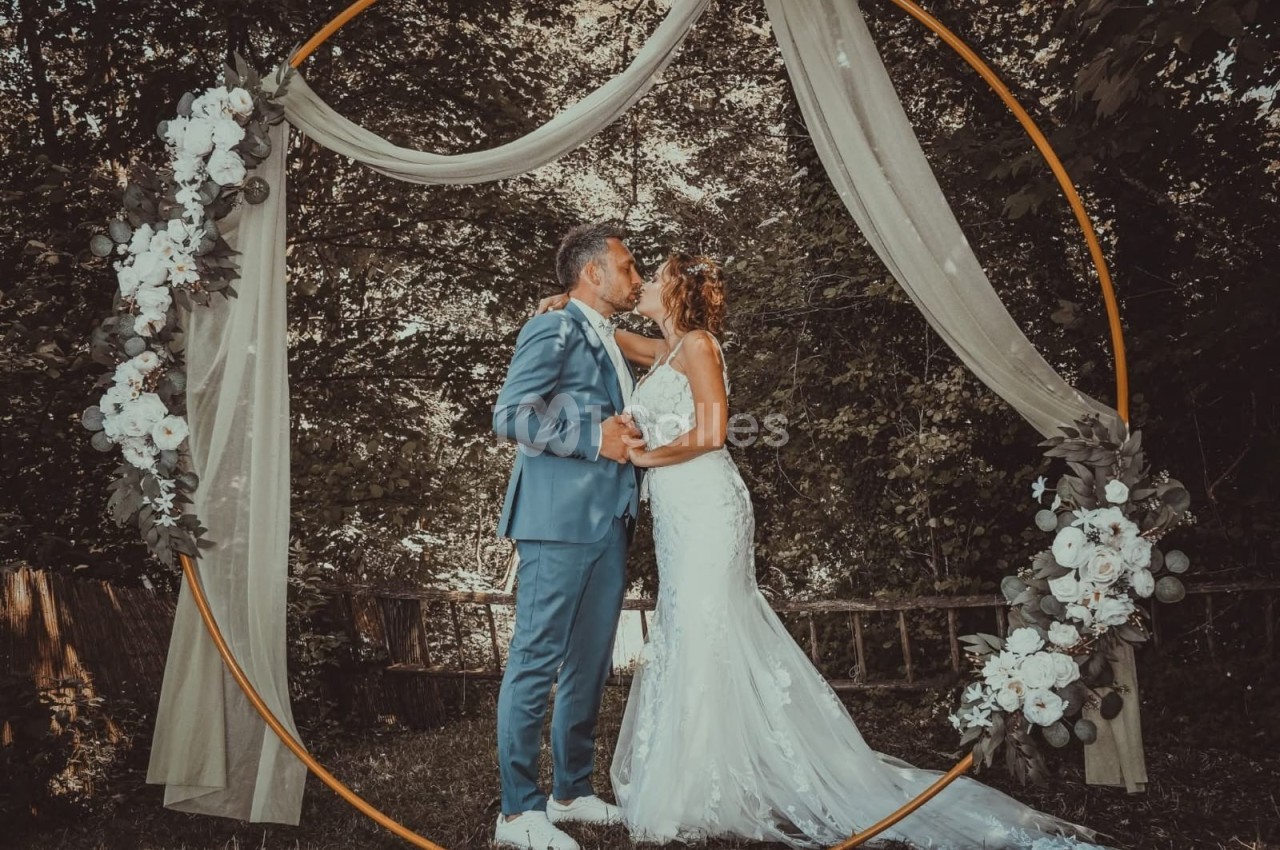 Un couple en tenue de mariage s'embrasse sous une arche décorée de fleurs dans un cadre naturel boisé.