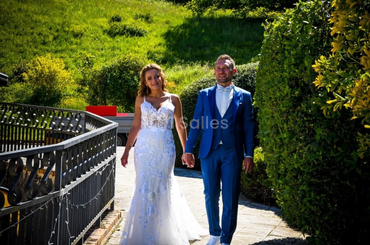 Un couple en tenue de mariage marche sur un chemin bordé de verdure par une journée ensoleillée.