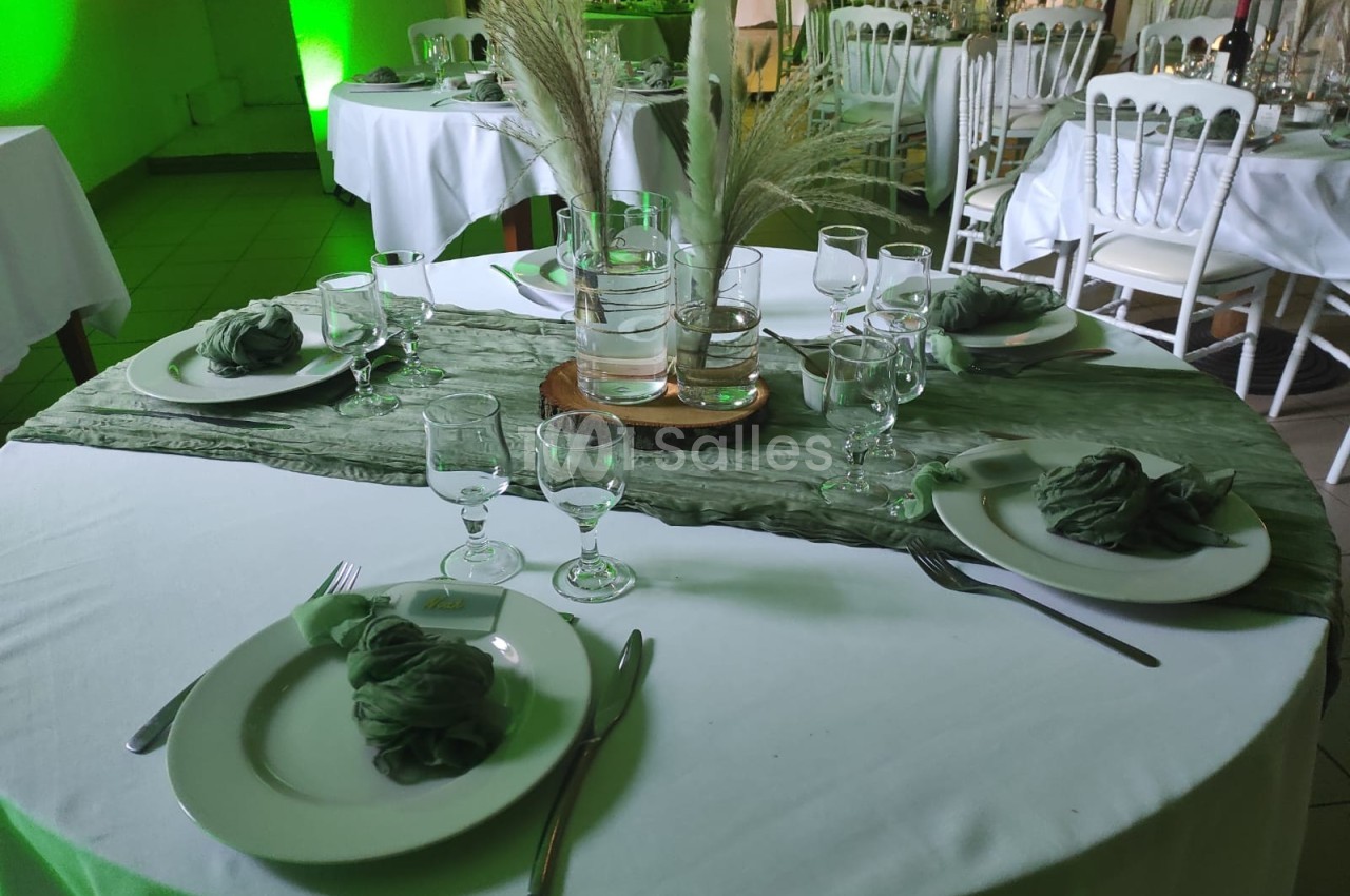 Table ronde dressée avec nappes blanches, serviettes vertes pliées, verres et centre de table avec vase et pampas.