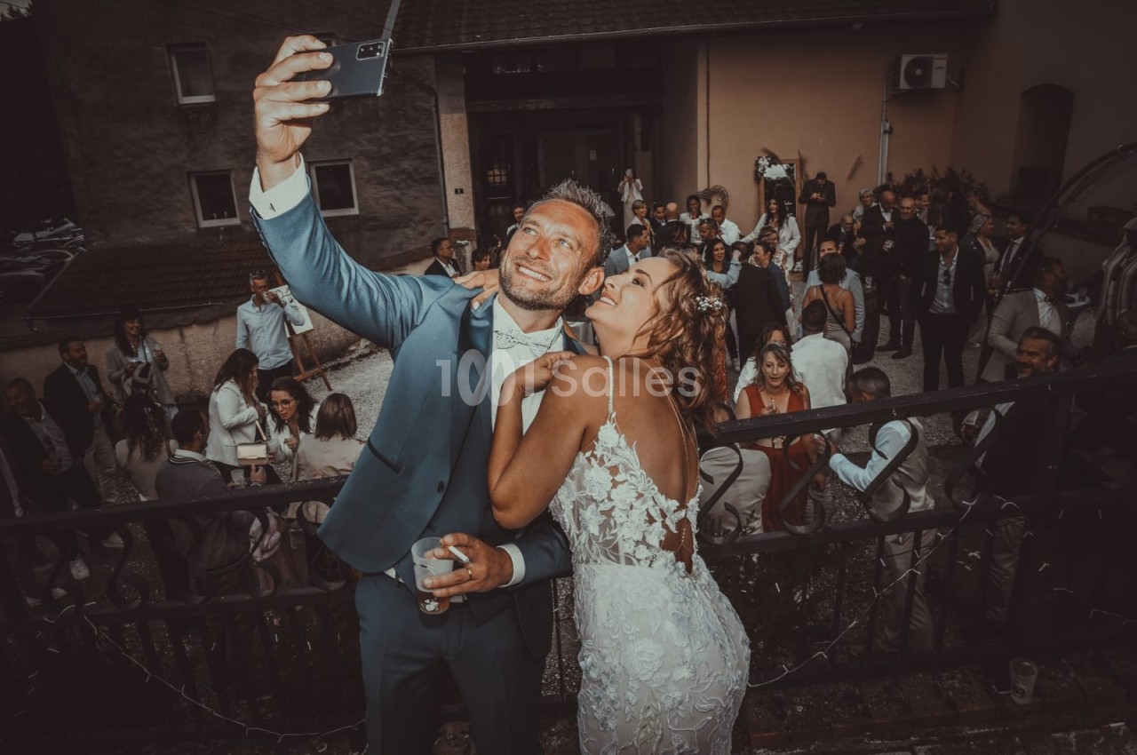 Un couple en tenue de mariage prend un selfie devant un groupe de personnes lors d'une réception en extérieur.