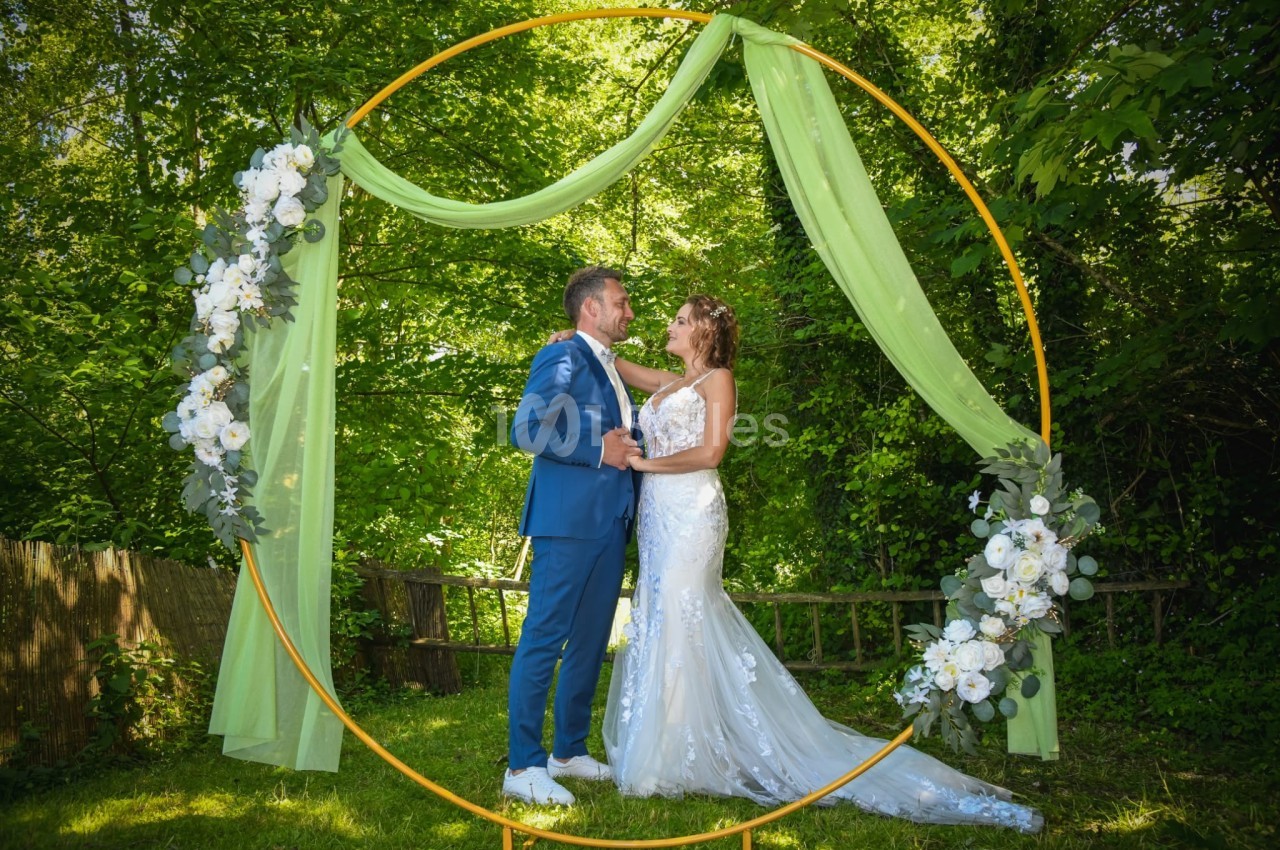 Un couple en tenue de mariage pose sous une arche circulaire décorée de fleurs blanches et de voiles verts, dans un jardin.