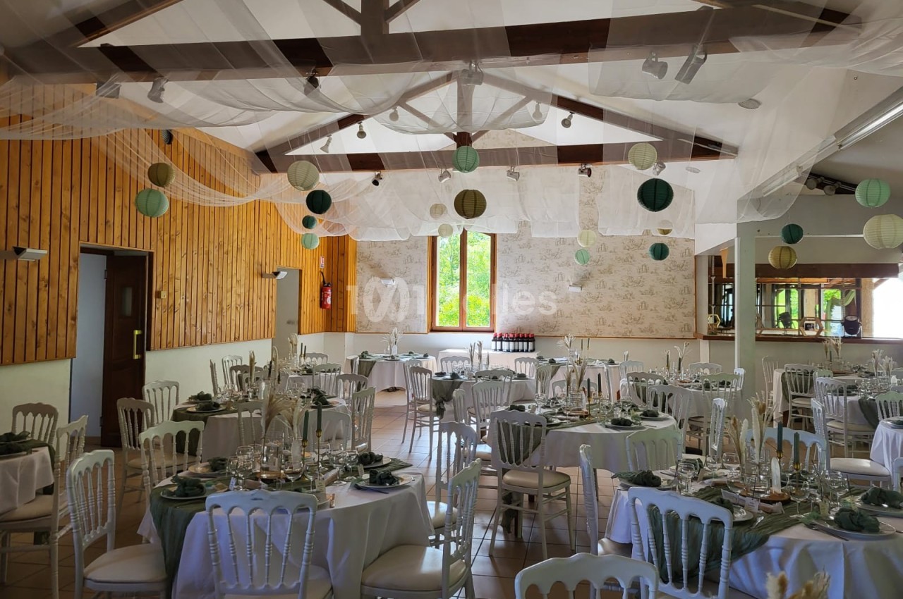 Salle décorée pour un événement avec tables rondes dressées, chaises blanches et lanternes suspendues au plafond.