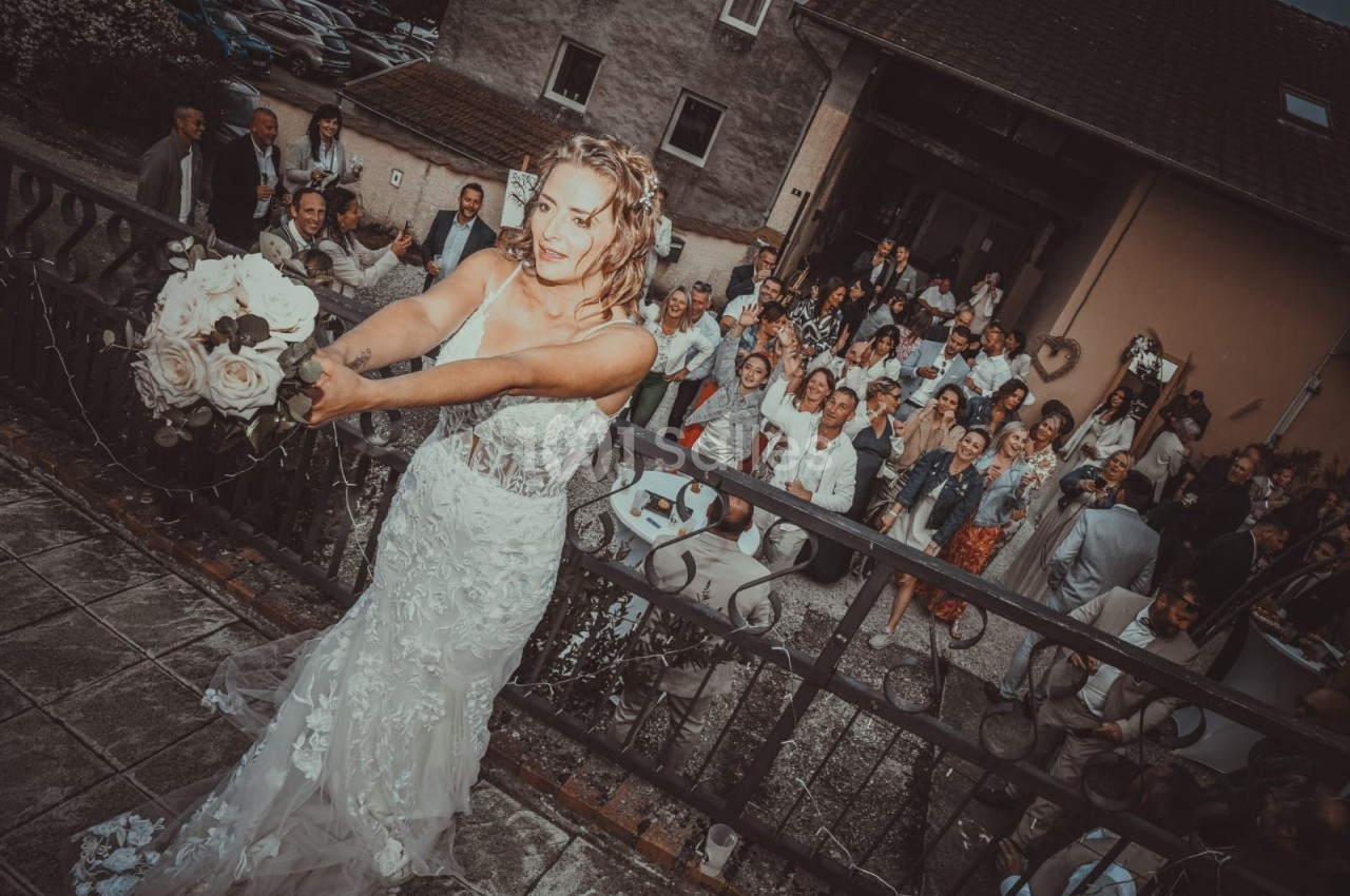 Une mariée lance son bouquet depuis un balcon devant un groupe d'invités rassemblés en contrebas.