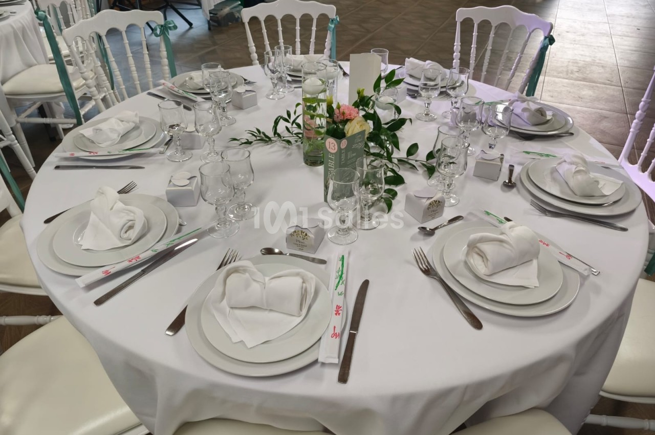 Table ronde élégamment dressée avec nappes blanches, vaisselle raffinée, serviettes pliées et décoration florale centrale.