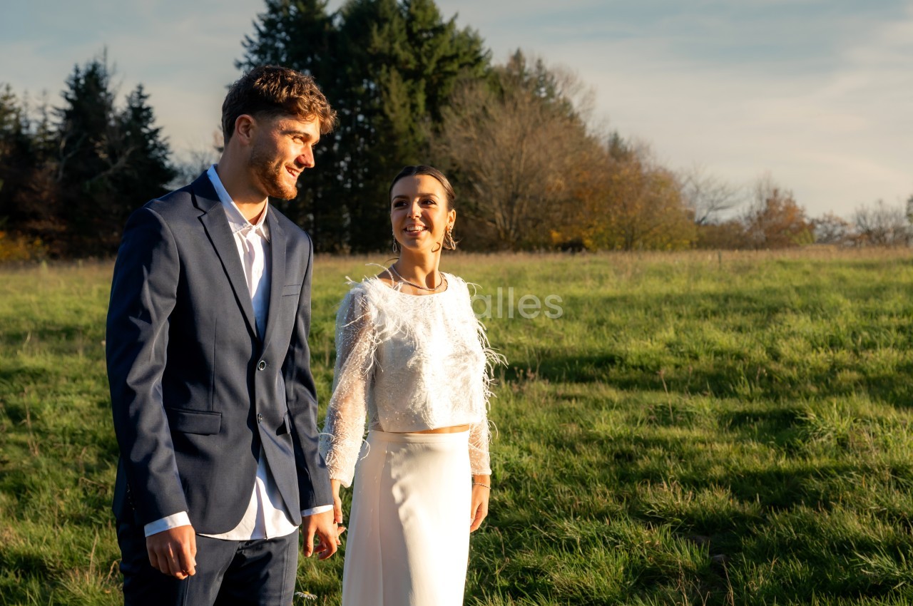 Un couple en tenue élégante marche dans un champ verdoyant sous une lumière douce de fin de journée.