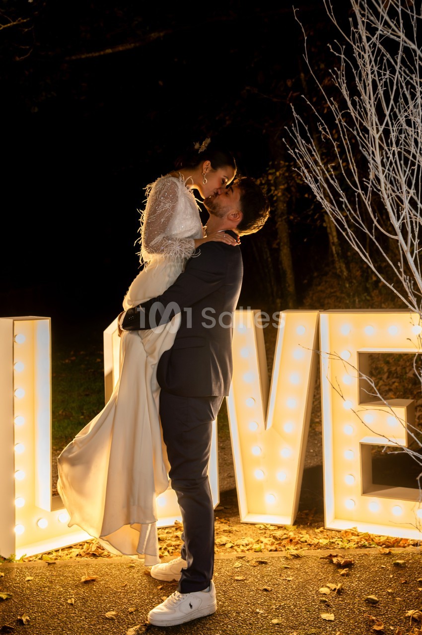 Un couple en tenue de mariage s'enlace devant des lettres lumineuses formant le mot ’LOVE’ dans un décor nocturne.
