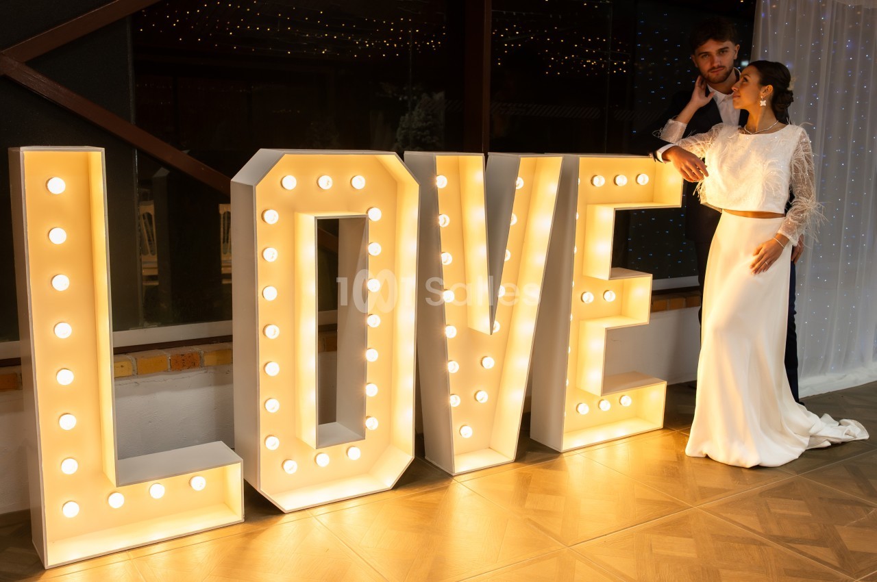 Un couple de mariés pose près de grandes lettres lumineuses formant le mot ’LOVE’ dans un décor festif.