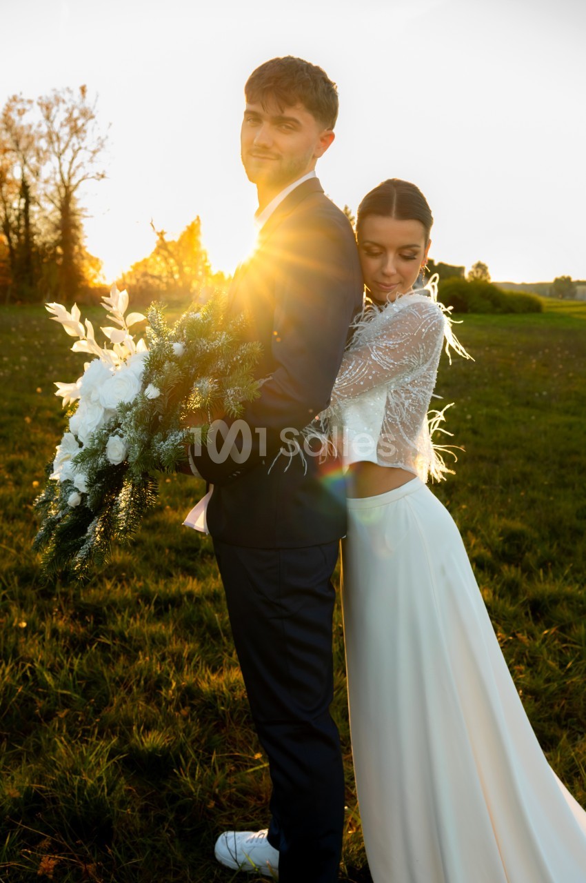 Un couple en tenue de mariage pose dans un champ au coucher du soleil, la mariée tenant un bouquet de fleurs blanches.