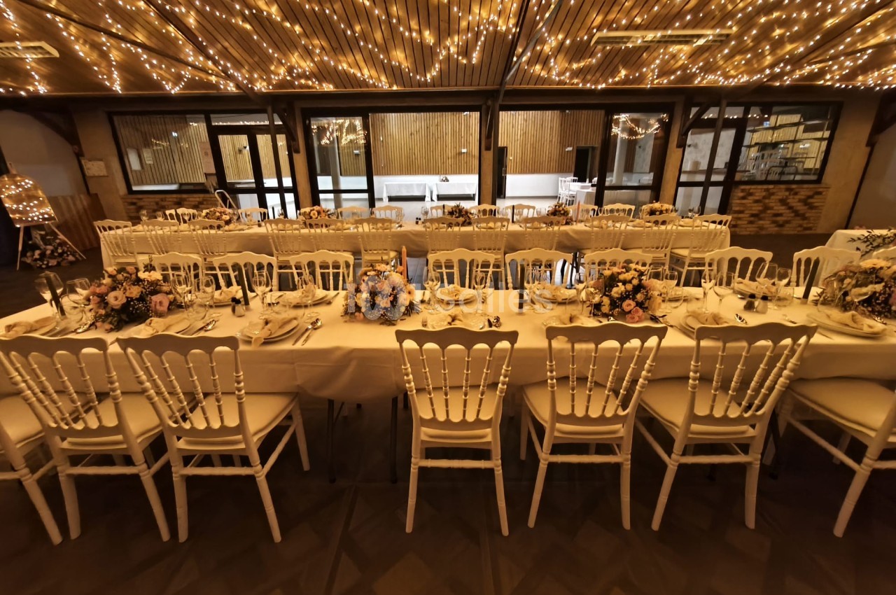 Salle de réception décorée avec des guirlandes lumineuses, tables dressées et chaises blanches alignées.