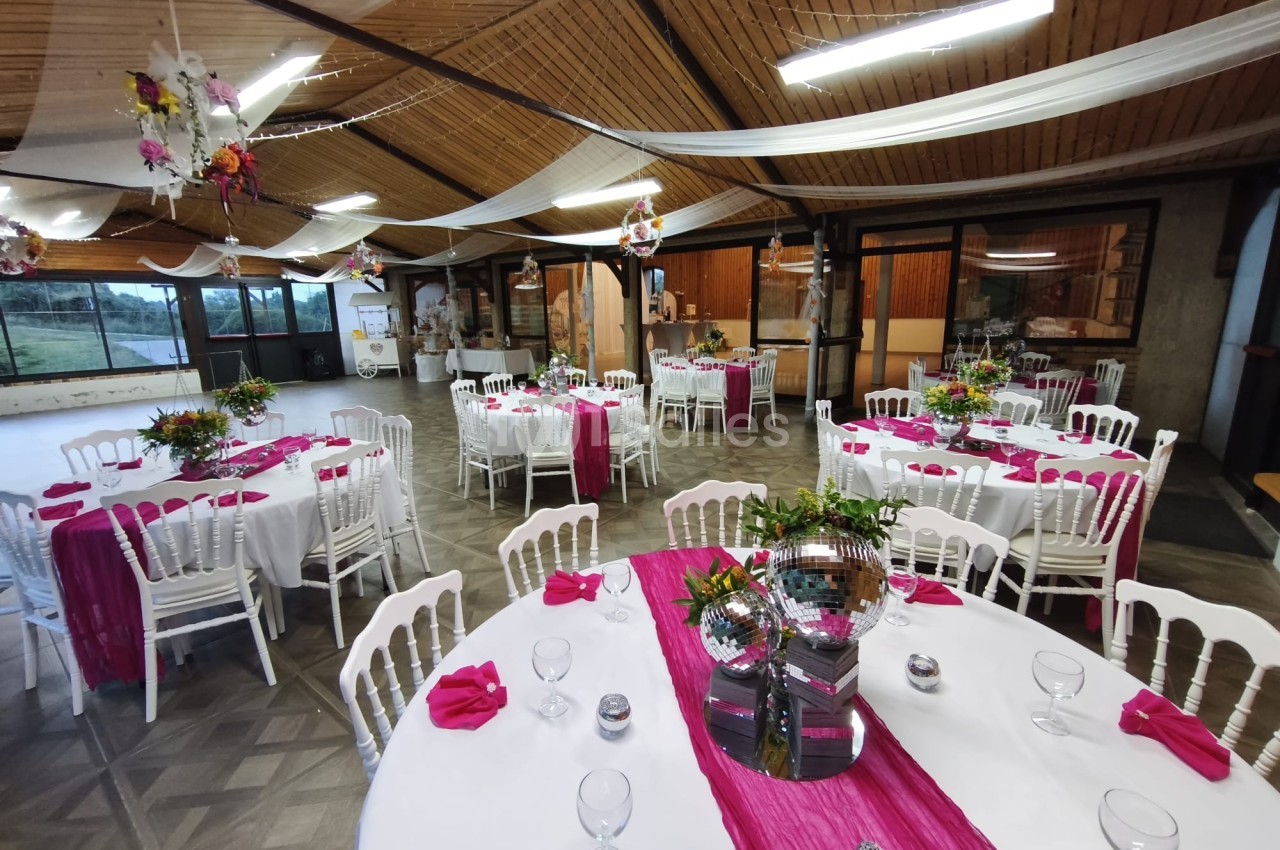 Salle de réception décorée avec des tables rondes, nappes blanches, chemins de table roses et chaises blanches.