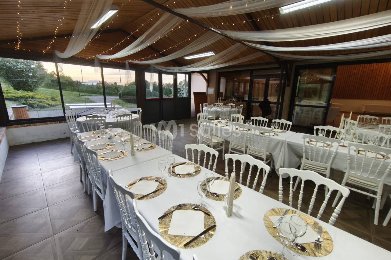 Salle de réception décorée avec des tables dressées, nappes blanches, chaises assorties et voilages suspendus.