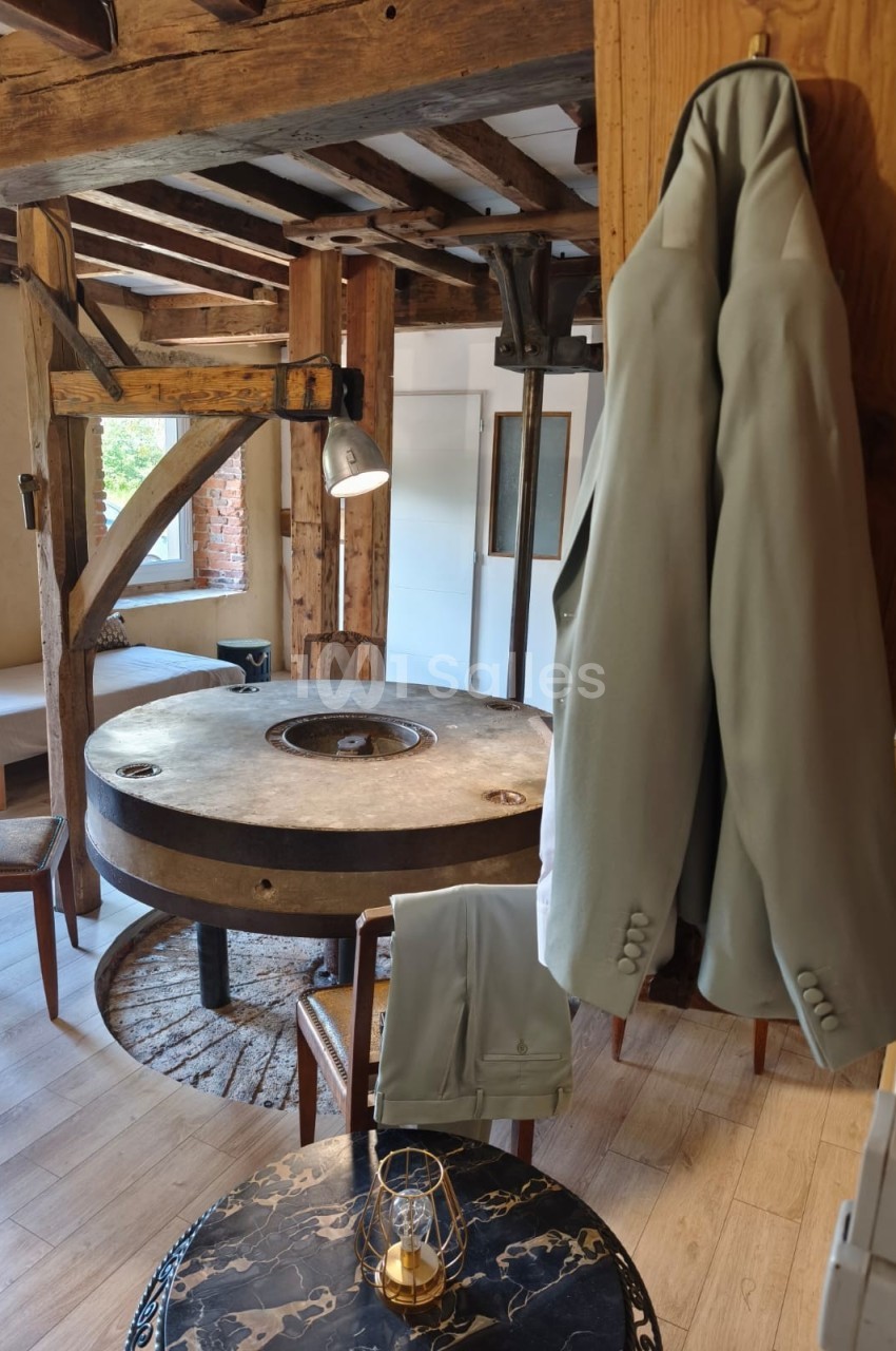 Deux vestes beiges suspendues dans une pièce rustique avec une table ronde en pierre et des poutres en bois apparentes.
