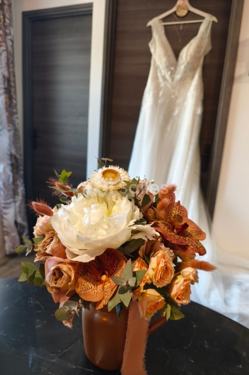 Bouquet de fleurs aux tons chauds posé sur une table, avec une robe de mariée suspendue en arrière-plan.