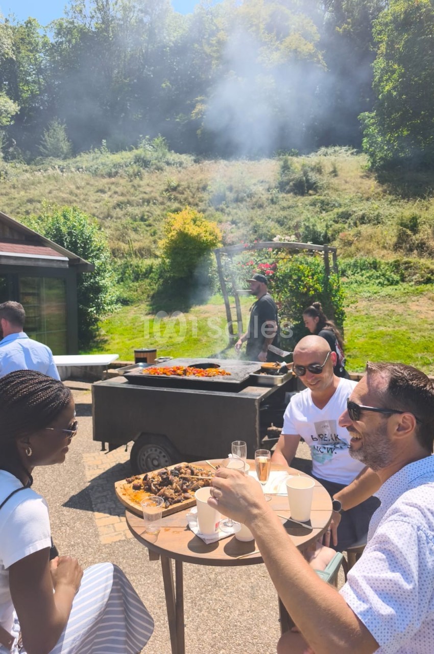 Des personnes discutent autour d'une table en extérieur près d'un barbecue en pleine cuisson, sous un ciel ensoleillé.