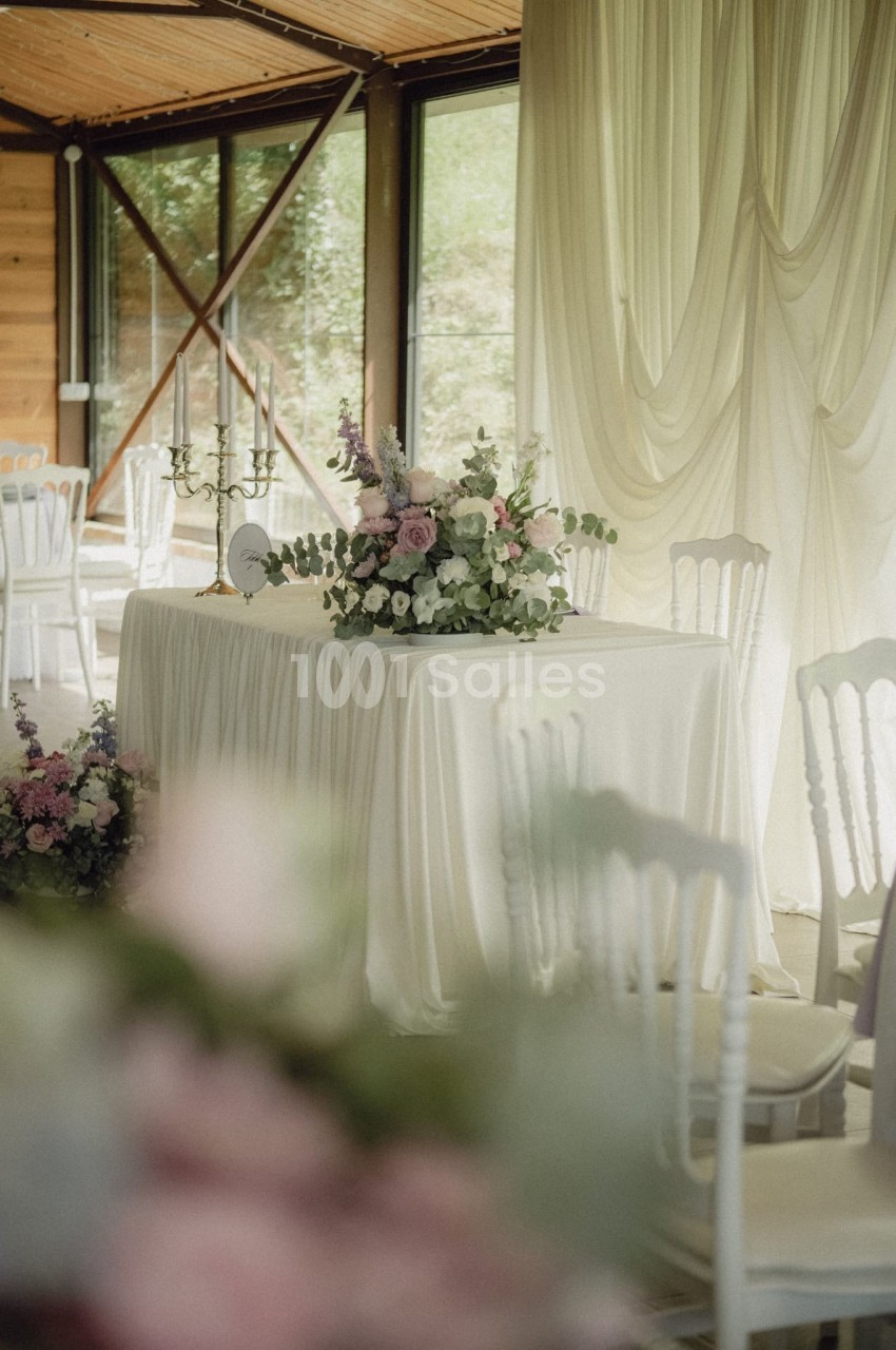 Table décorée avec une nappe blanche, un bouquet floral et des chandeliers, dans une salle lumineuse avec rideaux et baies…