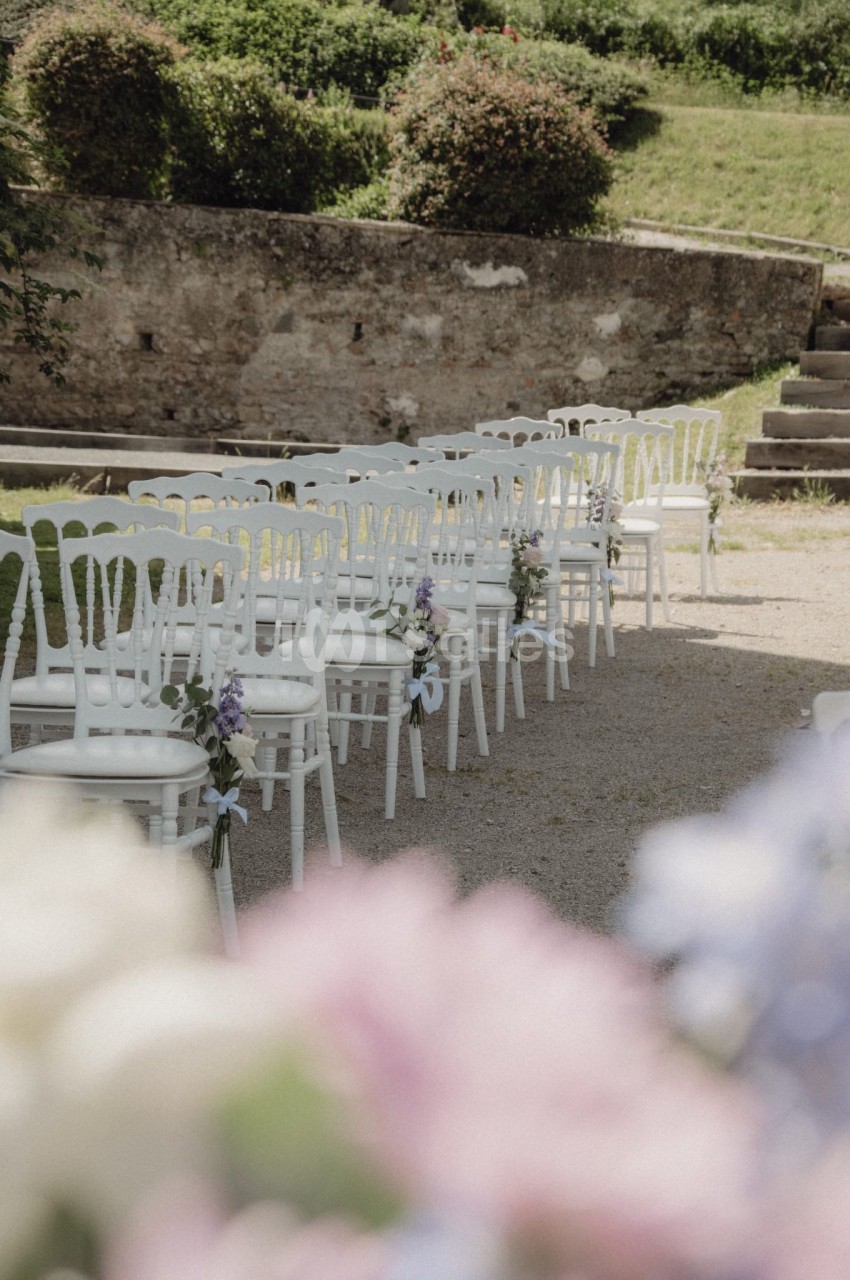 Chaises blanches alignées en extérieur, décorées de fleurs, sur un chemin gravillonné près d'un mur en pierre.