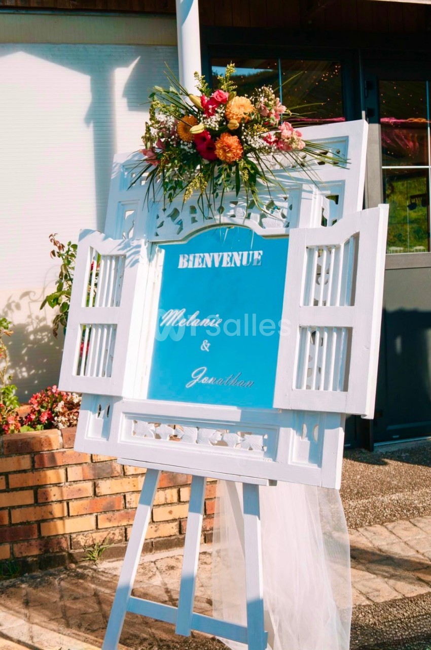 Panneau de bienvenue décoré de fleurs colorées, posé sur un chevalet blanc devant un bâtiment.
