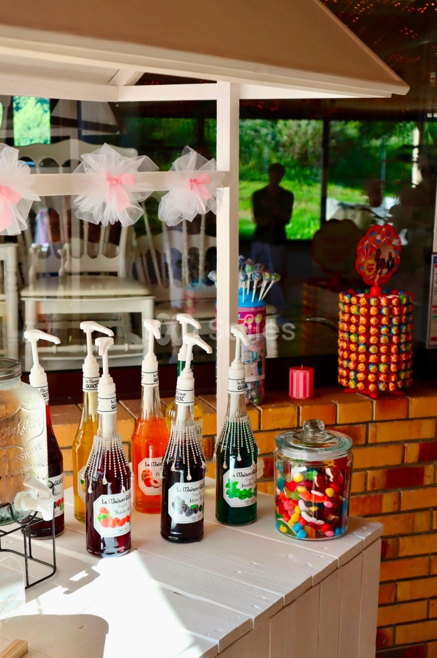 Stand avec sirops, bonbons colorés en bocaux et distributeur de chewing-gums, installé en extérieur lumineux.