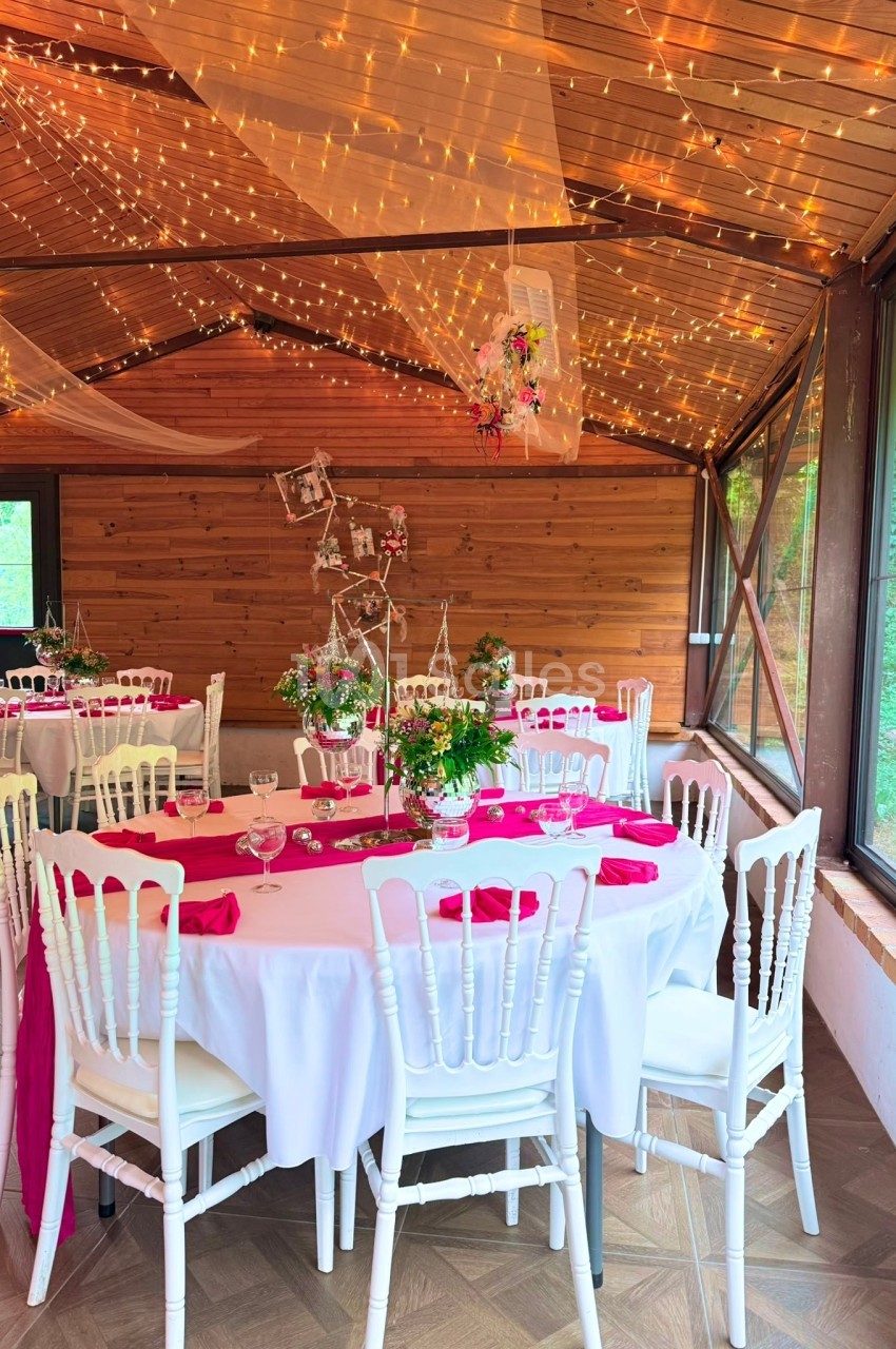Salle de réception en bois décorée avec des guirlandes lumineuses, tables rondes dressées avec nappes blanches et touches de…