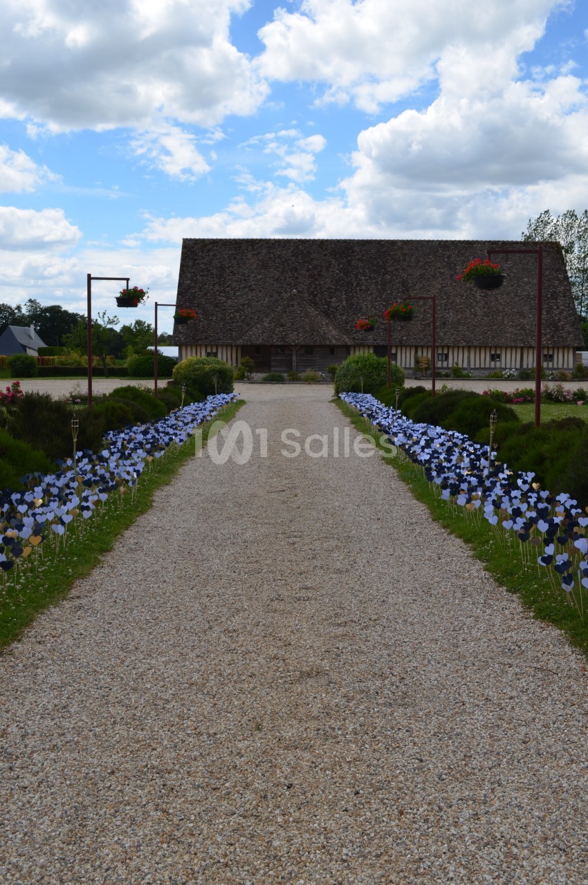 Allée gravillonnée bordée de fleurs décoratives menant à une maison à colombages sous un ciel partiellement nuageux.
