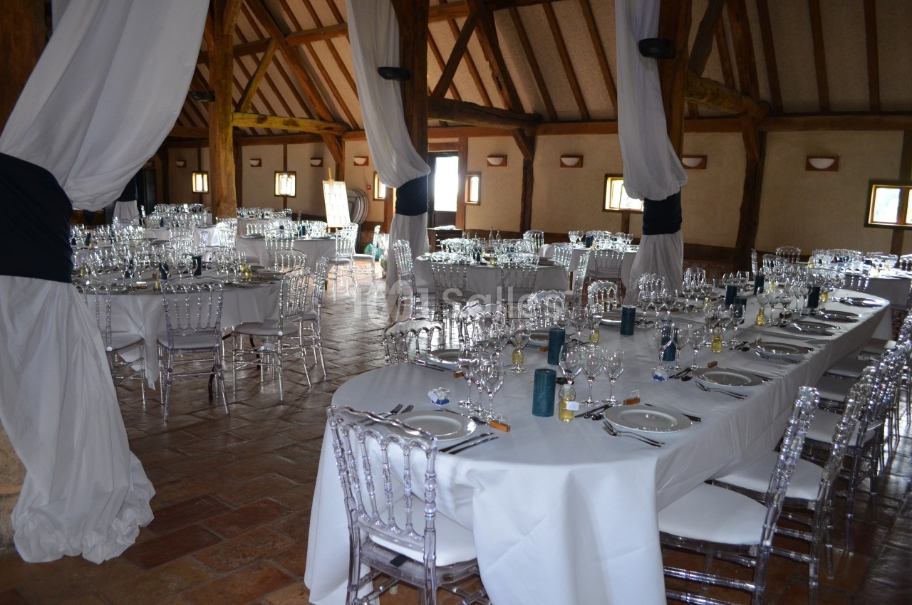 Salle de réception décorée avec des tables dressées, nappes blanches, chaises transparentes et poutres apparentes.