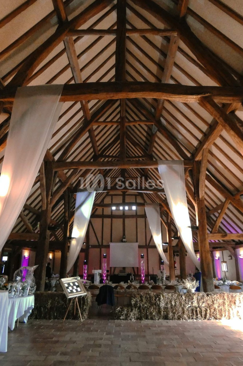 Grande salle de réception avec charpente en bois apparente, décorée de voilages blancs et éclairages colorés.