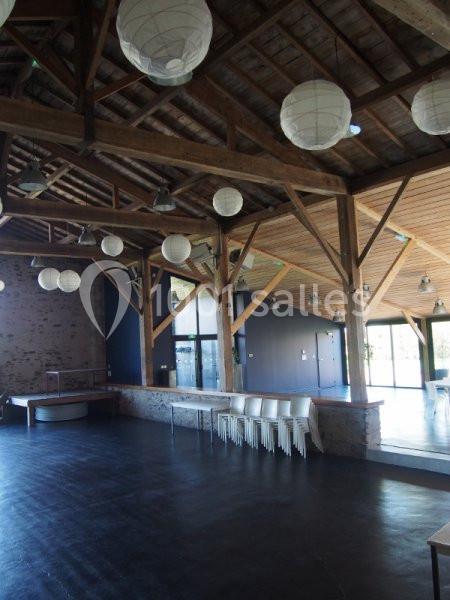 Salle spacieuse avec charpente en bois apparente, sol sombre, chaises empilées et lanternes suspendues.