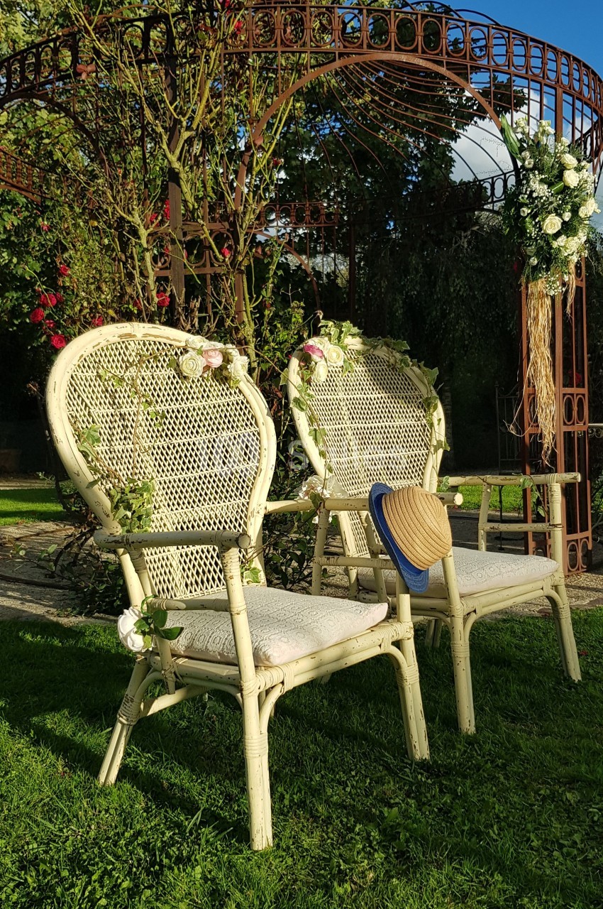 Deux chaises en osier décorées de fleurs, placées sur une pelouse devant une arche métallique ornée de végétation.