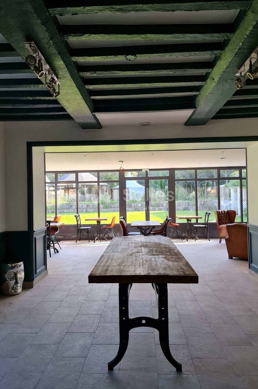 Vue intérieure d'une pièce lumineuse avec une grande table en bois, des poutres apparentes et des baies vitrées donnant sur…