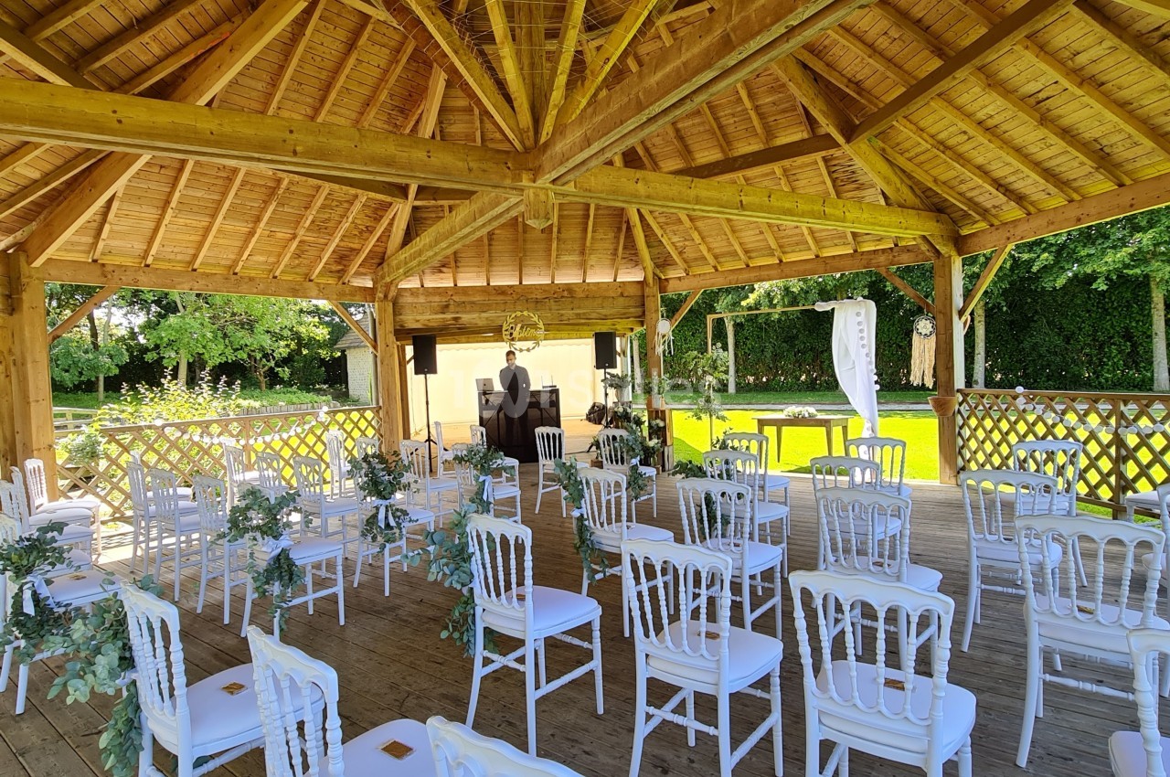 Chaises blanches disposées sous une structure en bois pour une cérémonie en extérieur, avec décoration florale.