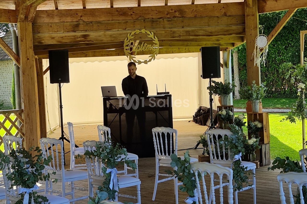 Un DJ se tient derrière sa table sous une pergola en bois décorée pour un événement avec des chaises blanches et des fleurs.