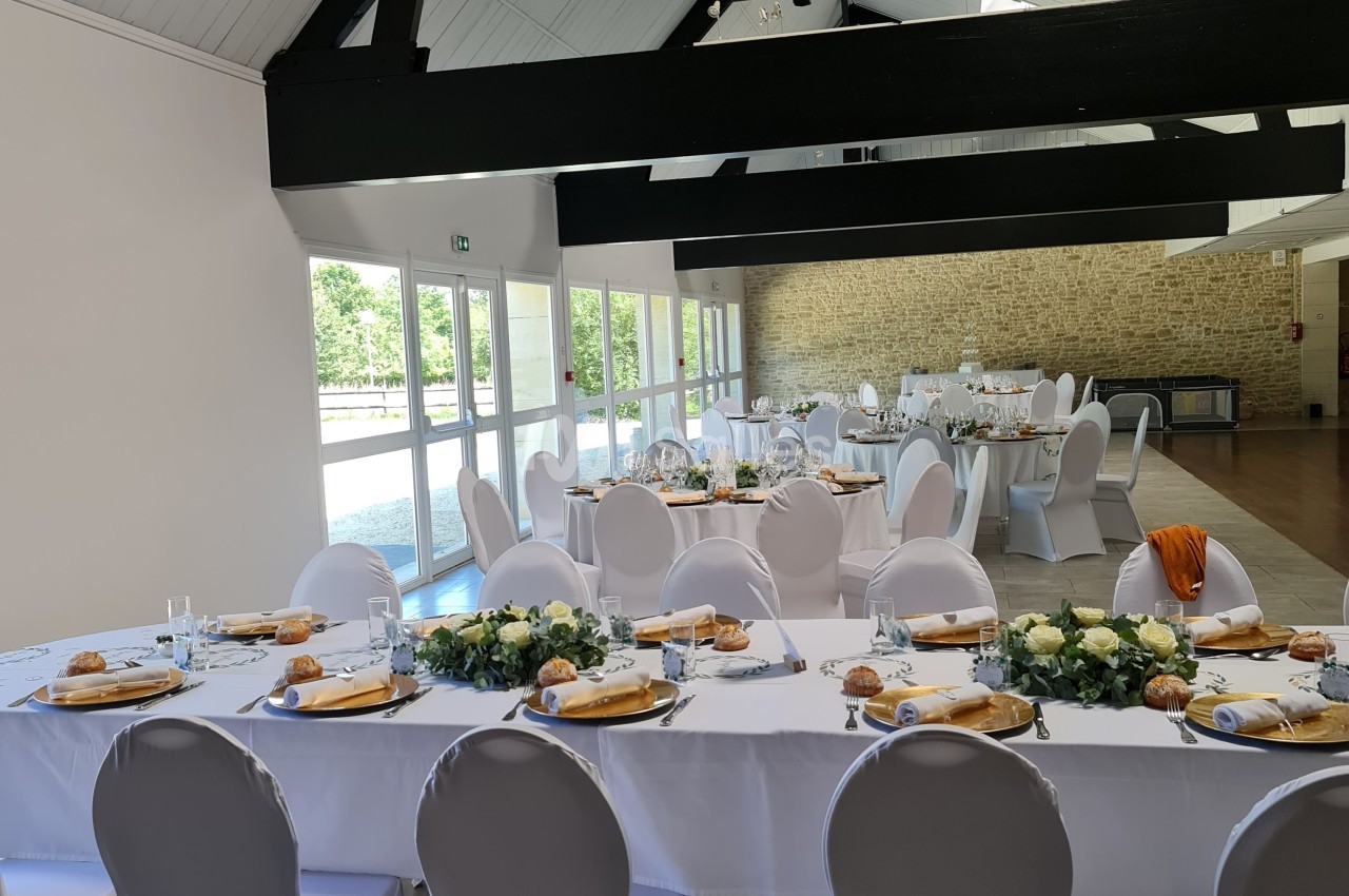 Salle de réception lumineuse avec tables décorées de nappes blanches, fleurs et vaisselle pour un événement.