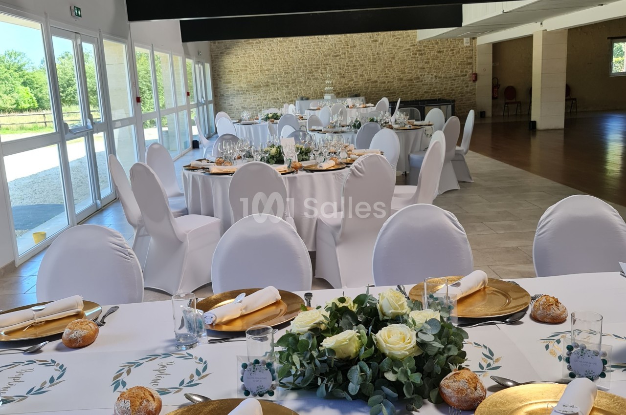Salle de réception lumineuse avec tables rondes dressées, nappes blanches, assiettes dorées et décoration florale.