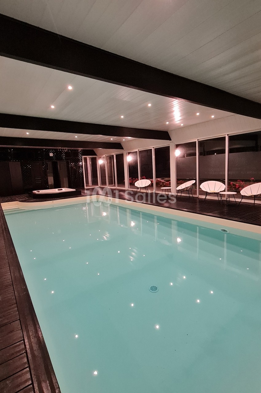 Piscine intérieure éclairée entourée de chaises modernes et de baies vitrées donnant sur un espace extérieur.