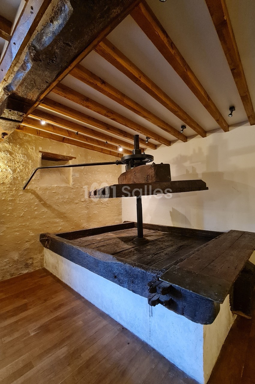 Pressoir ancien en bois dans une pièce avec murs en pierre, plafond à poutres apparentes et sol en parquet.