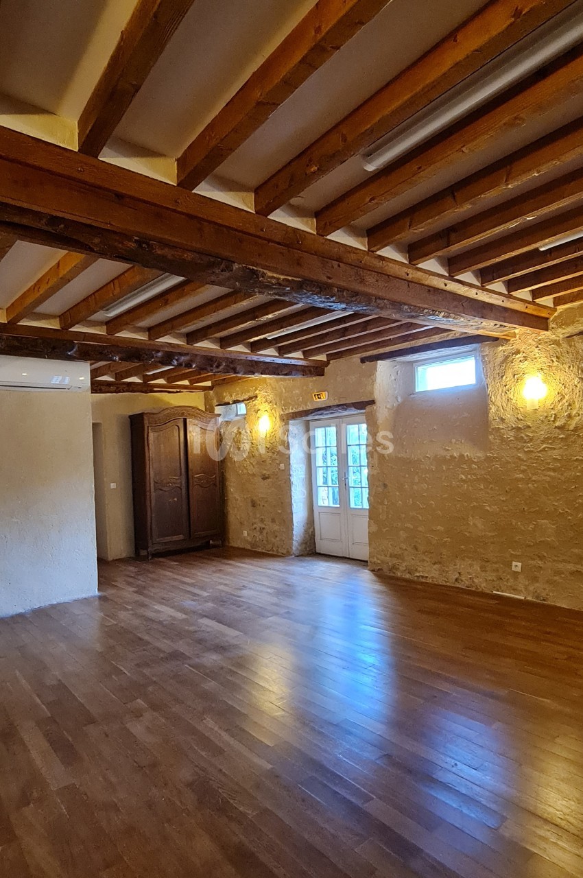 Salle avec parquet en bois, murs en pierre apparente, poutres au plafond, éclairage mural et porte-fenêtre vitrée.