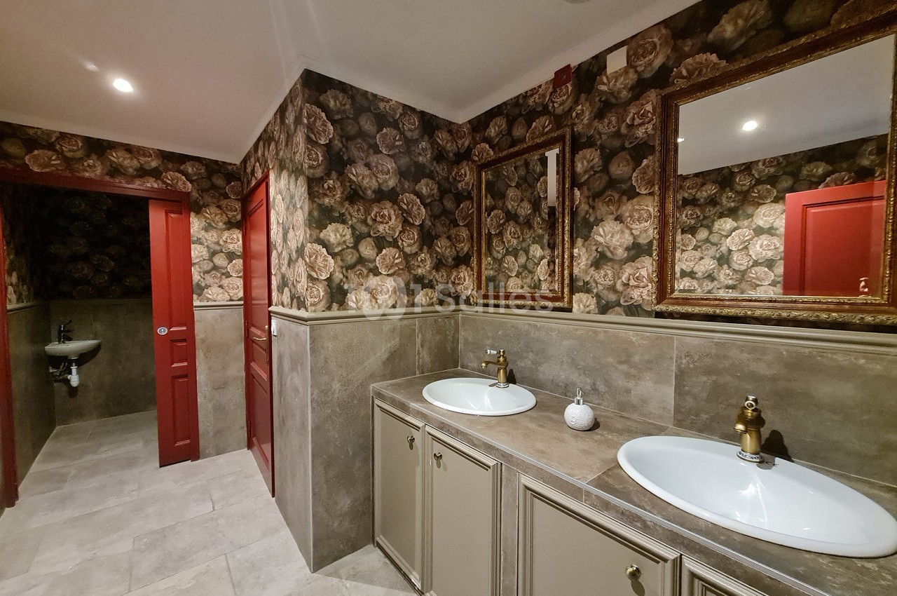 Salle de bain avec deux lavabos, miroirs dorés, murs à motifs floraux et portes rouges.