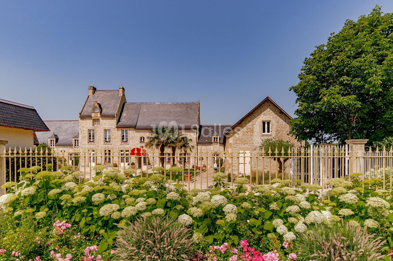 Façade d'une maison en pierre avec toit en ardoise, jardin fleuri au premier plan et ciel bleu en arrière-plan.