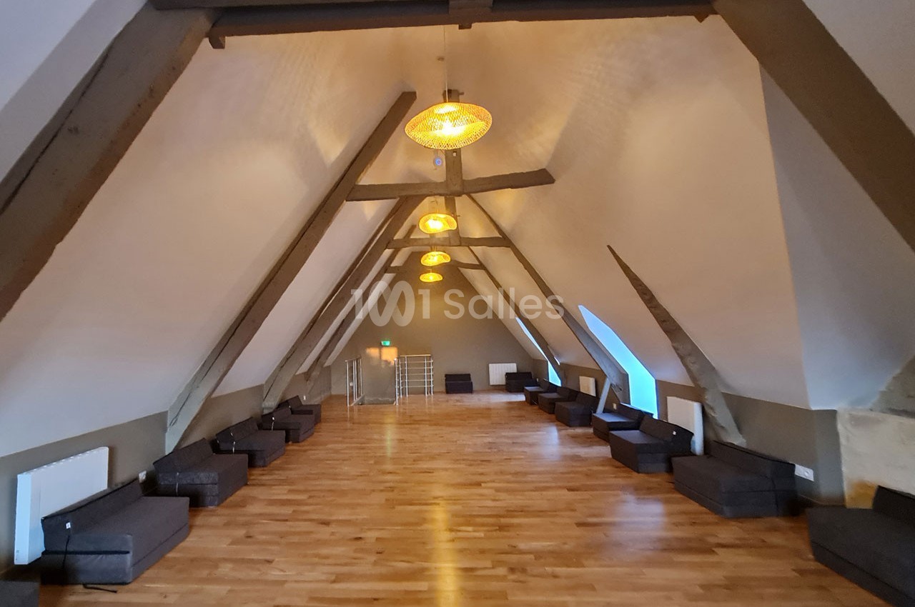 Grande salle mansardée avec poutres apparentes, parquet en bois clair, canapés alignés et éclairage suspendu.