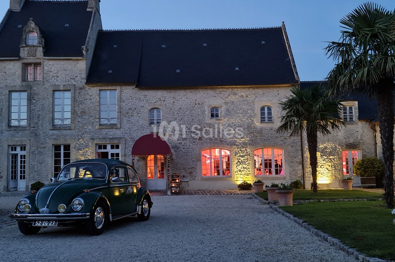 Voiture ancienne verte garée devant une grande bâtisse en pierre éclairée, avec palmiers et ciel au crépuscule.